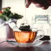 A hand is shown steeping a tea bag in a clear glass cup of tea. In the background, a vase of flowers and a glass jar are visible. Produktbild-Vorschau
