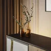 A close-up view of a dark wooden sideboard with two amber glass vases containing green branches. The background features brown curtains and a wall with vertical ribbed paneling in beige and gold. Produktbild-Vorschau