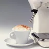 A close-up shot of a cream-colored espresso machine with a steaming wand positioned above a white cup of cappuccino, dusted with cinnamon. Podgląd zdjęcia produktu