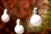 A string of white globe lights hangs in front of a blurred background of red and green foliage. Náhľad obrázku produktu