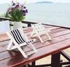 Two miniature deck chairs on a wooden table with a vase of flowers and the ocean in the background. Produktbild-Vorschau