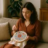 A woman is sitting on a couch and cross-stitching a floral pattern. Náhled obrázku produktu