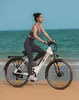 A woman is riding an electric bicycle on a sandy beach with the ocean in the background. Produktbild-Vorschau