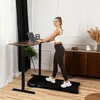 A woman is working on a laptop while standing on a treadmill next to a standing desk in a home office setting. Náhled obrázku produktu
