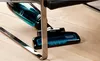 A cordless vacuum cleaner is shown on a wooden floor, partially underneath a chair. Náhľad obrázku produktu