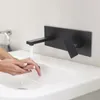 A person is washing their hands under a black wall-mounted faucet in a white sink. Podgląd zdjęcia produktu