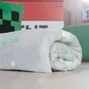 A rolled-up blanket with a Minecraft Creeper pattern is shown on a wooden floor, with a pillow and a red storage box in the background. Podgląd zdjęcia produktu