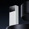 A white power bank with a black digital display is shown against a dark background, with black circular elements framing it. Produktbild-Vorschau