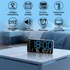 A digital clock is shown on a table in front of a blurred bedroom background. Above the clock, there are icons and text describing its features. Produktbild-Vorschau