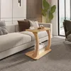 A wooden C-shaped side table is placed next to a cream-colored sofa in a living room. The table has a shelf with books and holds two cups and pastries. Náhled obrázku produktu