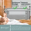A baby is sitting in a crib next to a bed with blankets. Produktbild-Vorschau