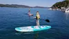 Two children are paddleboarding on a lake on a sunny day. There are boats and houses in the background. Náhled obrázku produktu