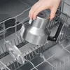 A hand is placing a stainless steel kitchen appliance into a dishwasher rack. A clear plastic lid is also in the rack. Produktbild-Vorschau