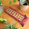 A wooden Kalaha board game with colorful stones is placed on a wooden table, surrounded by houseplants. Produktbild-Vorschau