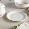 A white ceramic saucer is placed on a light-colored tablecloth, with a sugar bowl and a bowl with a spoon visible in the background. Produktbild-Vorschau