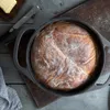 A rustic loaf of bread in a cast iron pot on a wooden surface, with butter and a knife nearby. Produktbild-Vorschau