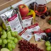 A collection of wine yeast nutrient packets and fresh fruit arranged on a wooden surface, with a glass of wine in the background. Produktbild-Vorschau