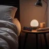 A digital alarm clock is placed on a nightstand next to a bed. Produktbild-Vorschau