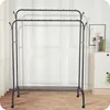 A black metal garment rack with two tiers and a shoe rack at the bottom, set against a white wall with decorative molding. A small cabinet with books and a framed picture is visible to the right. Anteprima dell’immagine prodotto