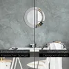 A split image showing two different styles of a round mirror mounted on a textured gray wall above a desk. The left side shows a mirror with a simple frame, and the right side shows a mirror with a decorative, colorful frame. Produktbild-Vorschau