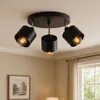 A black ceiling light fixture with three adjustable spotlights is shown mounted on a white ceiling. The background includes a wall with a framed picture and a window with curtains. Podgląd zdjęcia produktu