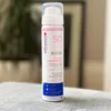 A white spray bottle of Ultrasun SPF 50 Face sunscreen is placed on a textured gray surface. Podgląd zdjęcia produktu