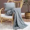 A gray blanket and pillow are draped over a rattan chair, with a plant in a basket in the background. Podgląd zdjęcia produktu