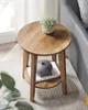 A round wooden side table with a lower shelf is placed next to a sofa, with a potted plant on top and a decorative vase and book on the shelf. Produktbild-Vorschau