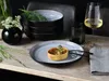 A table setting with a quiche on a plate, a stack of bowls, a glass of wine, and a vase of flowers. Náhľad obrázku produktu