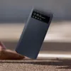 A dark gray Samsung flip case for a phone is shown falling onto a textured surface. Náhled obrázku produktu