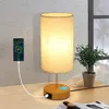 A bedside lamp with a phone charging on it, placed on a wooden desk next to a plant. Produktbild-Vorschau