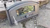 A cast iron oven door with a glass window rests on a stone and gravel surface. Produktbild-Vorschau