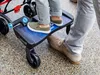 A child stands on a buggy board attached to a stroller, while an adult's legs are visible next to it. Produktbild-Vorschau