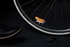 A small, orange and white hamster-shaped object is attached to the spokes of a bicycle wheel. A black shoe is visible in the foreground, and several dark stones are scattered on the dark surface. Podgląd zdjęcia produktu