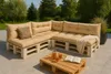 An outdoor patio setting with a sectional sofa made of pallets and beige cushions, a wooden pallet coffee table with drinks and a book. Anteprima dell’immagine prodotto
