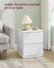 A white nightstand with a lamp and a cup on top, next to a sofa in a room. Produktbild-Vorschau