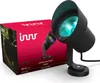 A product box for an Innr outdoor spotlight is shown next to the spotlight itself. The spotlight is black and emits a blue light. The box is red and features the Innr logo and an image of a garden path at night. Produktbild-Vorschau