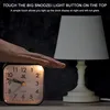 A hand is touching the top of an analog alarm clock, which is next to a table lamp. Produktbild-Vorschau