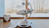 A baby is sitting in a LIONELO Bella electric baby bouncer in a living room with a large window showing a snowy landscape. Produktbild-Vorschau
