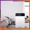 An air purifier is placed on the floor next to a bed in a bedroom, with a nightstand and a window in the background. Podgląd zdjęcia produktu
