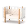 A white baby crib with wooden accents and a light-colored mattress and bumper pad, shown against a white background. Produktbild-Vorschau