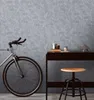 A bicycle is parked next to a desk with decorative items and a stool in front of a textured gray wall. Produktbild-Vorschau