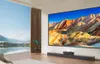 A modern living room with a large projector screen displaying a colorful mountain landscape, a projector on a media console, and a large window with a view of a lake and mountains. Produktbild-Vorschau