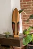 A decorative wooden surfboard on a shelf next to a plant and a brick wall. Produktbild-Vorschau