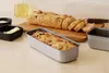 A loaf of bread is sliced on a wooden cutting board, with a loaf pan containing unbaked bread in the foreground, and small dishes of salt and butter to the left. Voir les images du produit