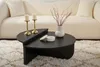 A modern black coffee table with a semicircular design sits on a fluffy cream rug in front of a white couch. Two brass candlesticks with white candles and a white vase with dried branches are on the table. Produktbild-Vorschau