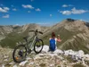 A person with a backpack and hiking poles sits on a rocky outcrop overlooking a mountain valley, with a bicycle parked next to them. Produktbild-Vorschau