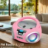 A pink boombox with a CD player and LED lights sits on a wooden table in a living room with large windows overlooking a lake and trees. Voir les images du produit