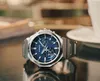 A watch is placed on a reflective surface, with a blurred background of a cafe setting. Produktbild-Vorschau