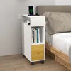 A narrow white bedside table with a wood-tone drawer and shelves, placed next to a bed. A phone and smartwatch are charging on top. Voir les images du produit